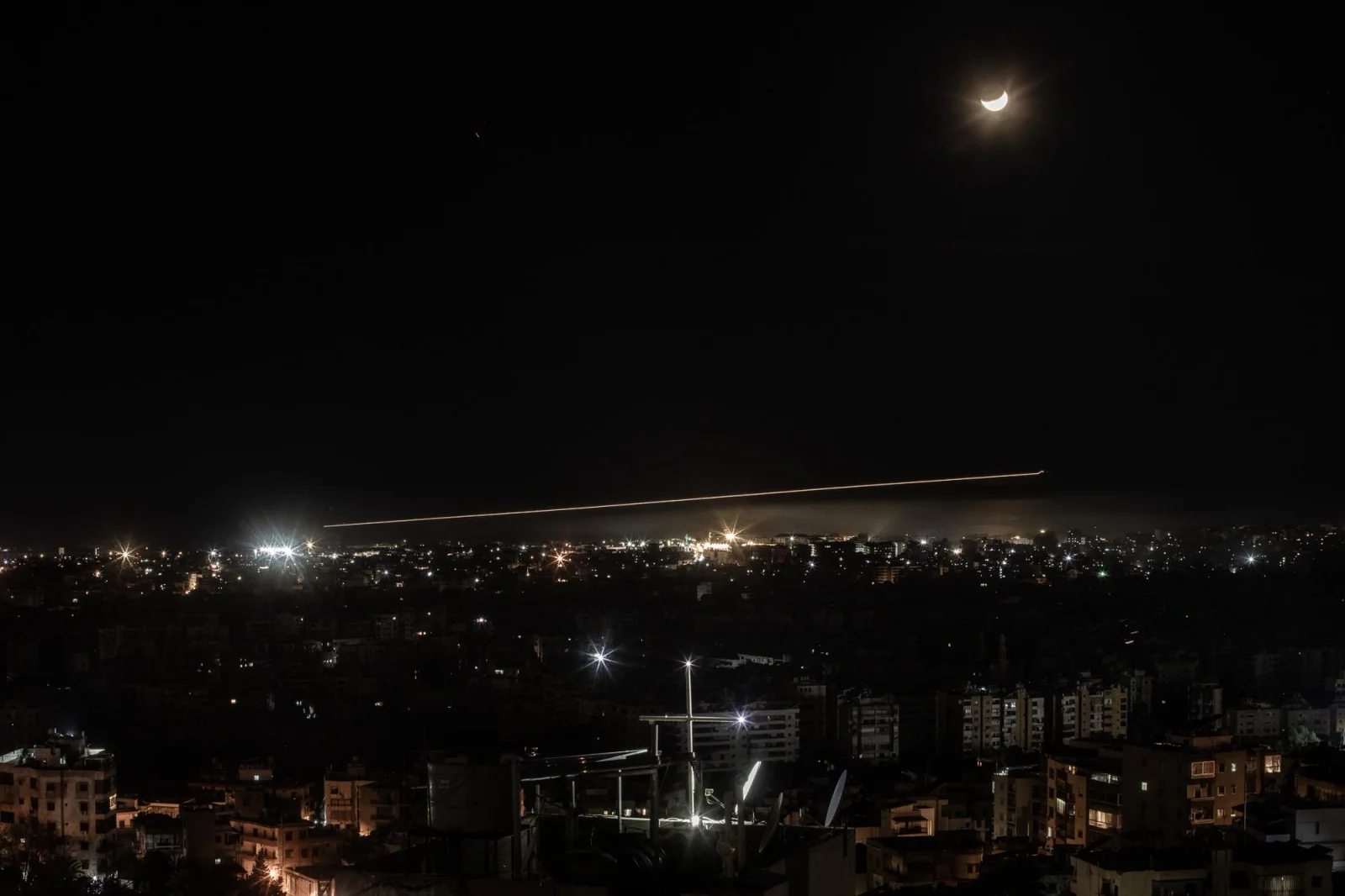 El skyline de Dahie, los suburbios del sur de Beirut, se mantiene a oscuras y bajo la amenaza constante de los bombardeos israelíes, que se concentran en esta zona densamente poblada. Al fondo, un avión de la compañía Middle East Airlines (MEA) se aproxima a la pista de aterrizaje del Aeropuerto Internacional Rafic Hariri. Líbano